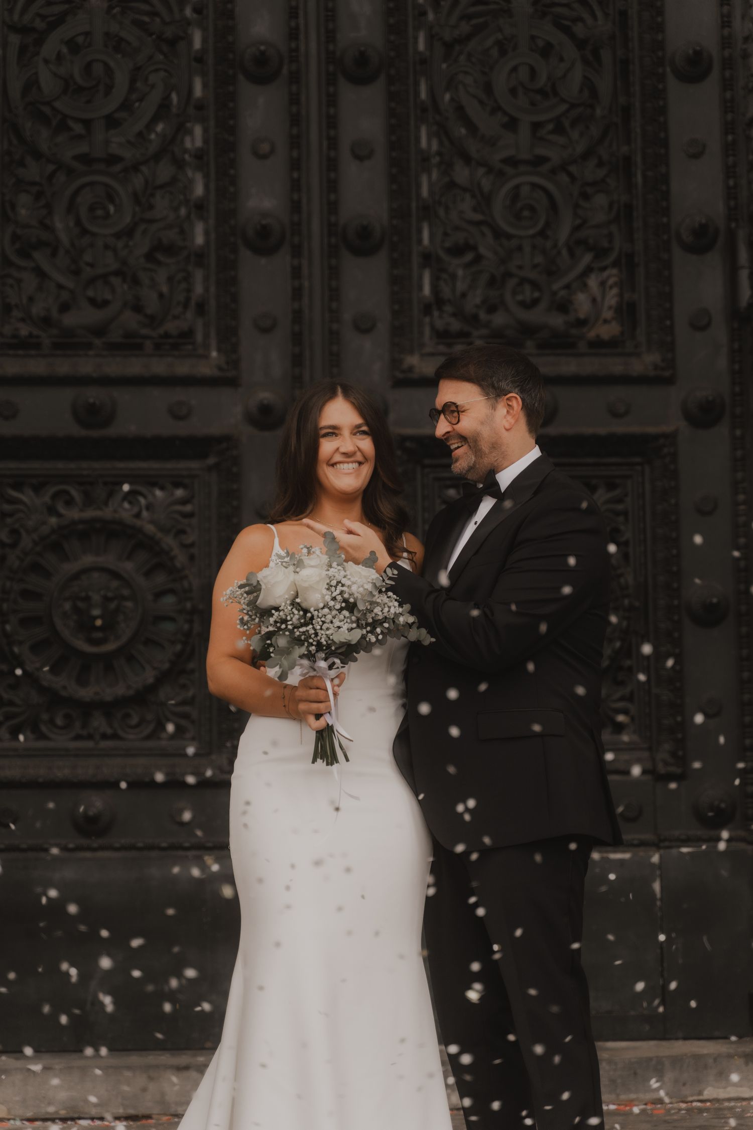 A bride and groom share a joyful moment in front of ornate church doors while confetti falls around them.