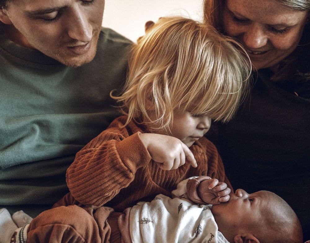 A young child in a brown sweater gently touches a baby while two adults watch closely.