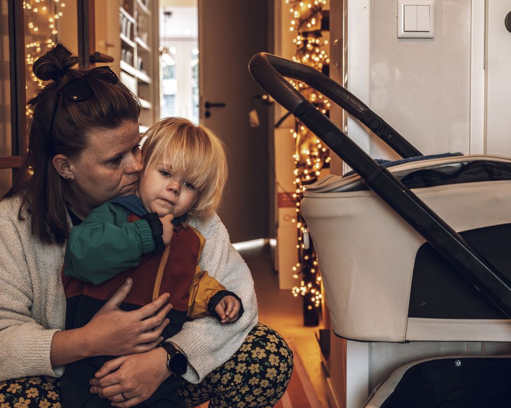 A cozy holiday scene with a child in a green sweater being held near twinkling Christmas lights and a stroller.