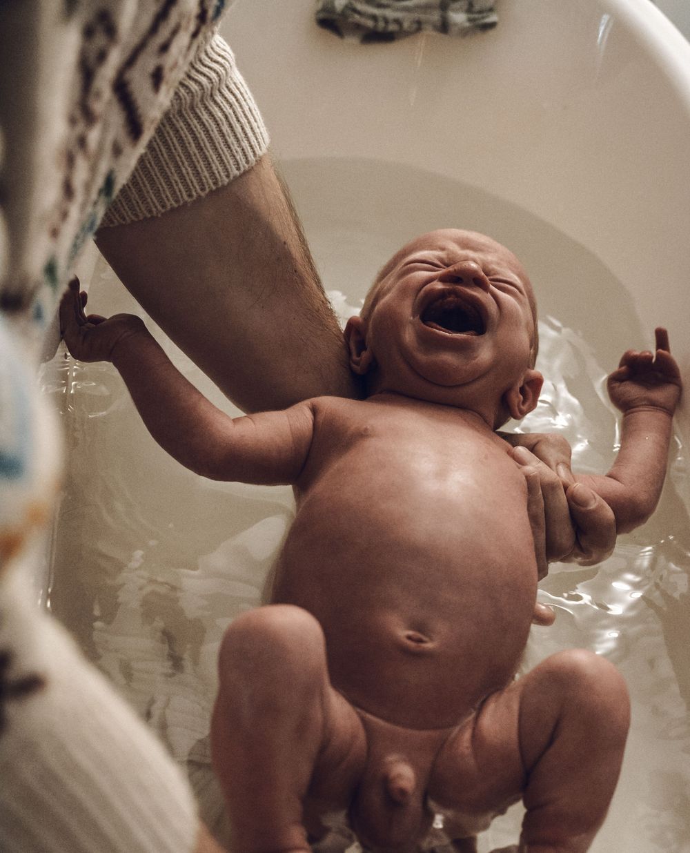 A laughing baby splashes during bath time in a white bathtub.