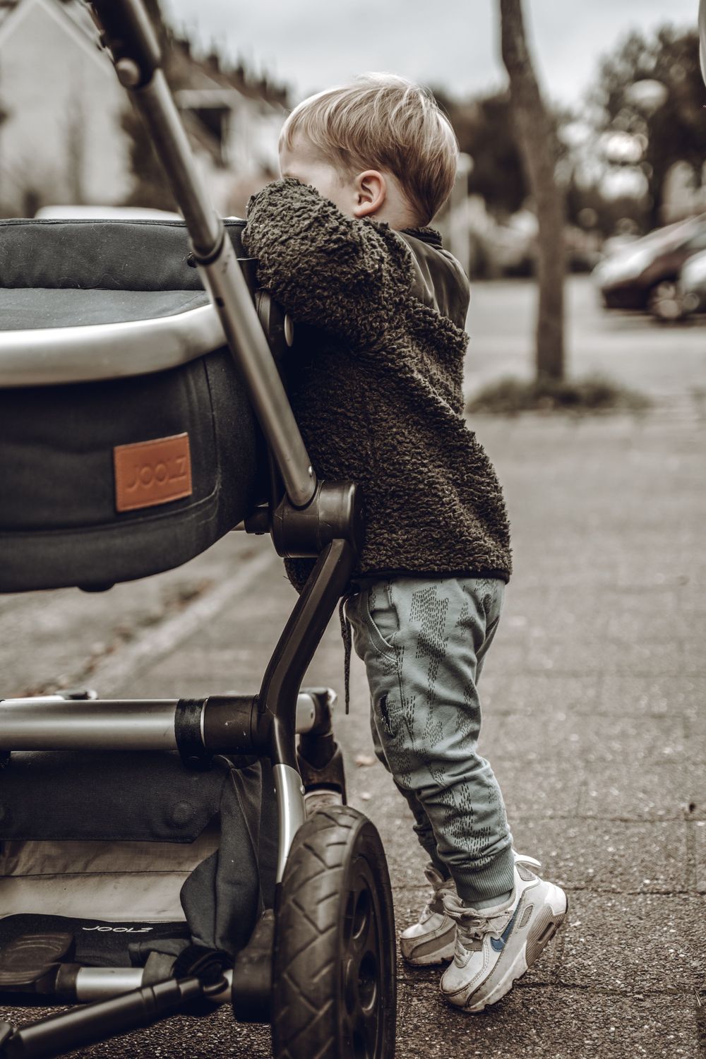 A small child in a brown coat and jeans reaches up to touch the wheel of a stroller on a paved path.