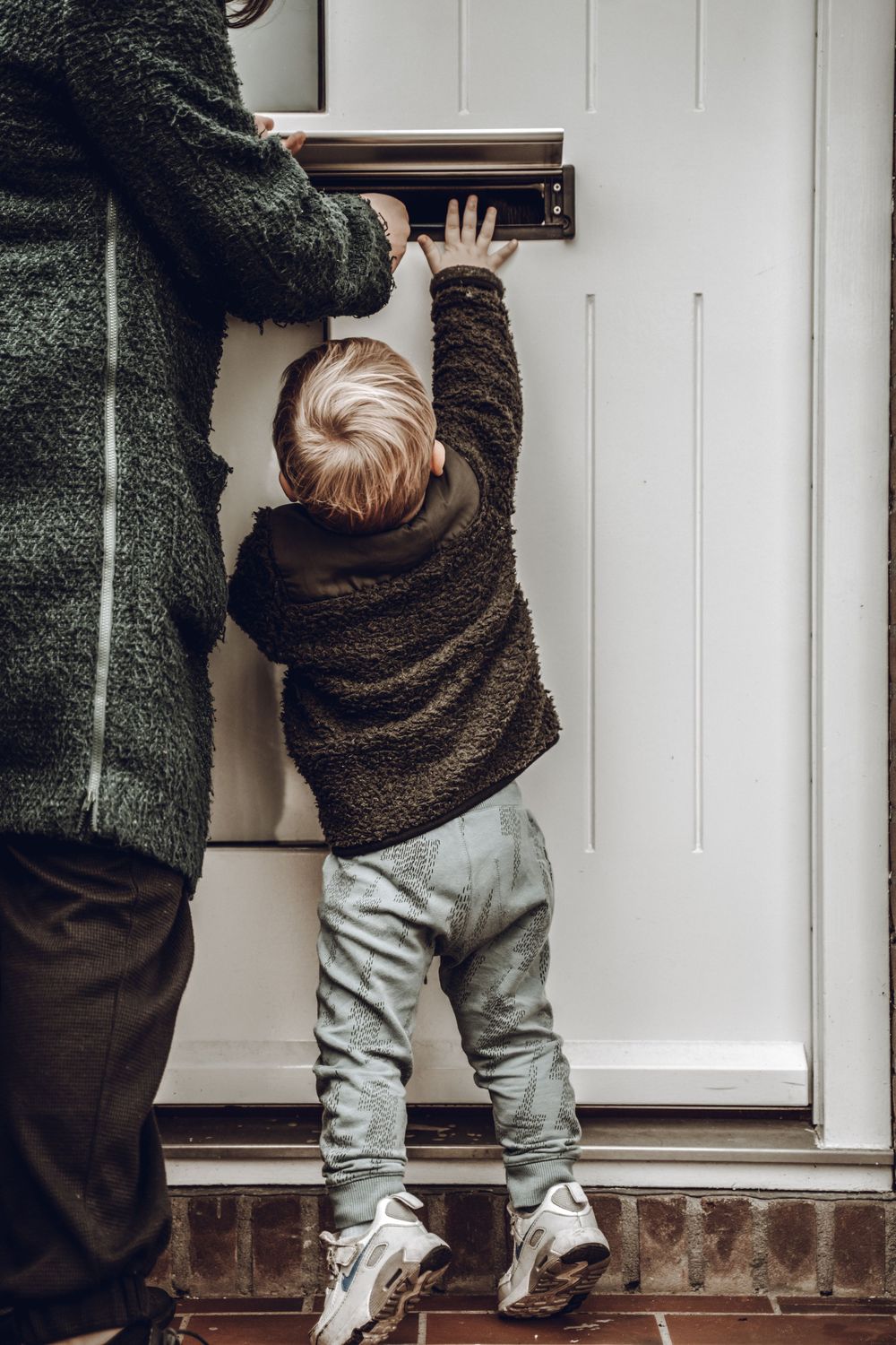 A small child in gray pants and white shoes reaches up to open a white door while wearing a brown fuzzy sweater.