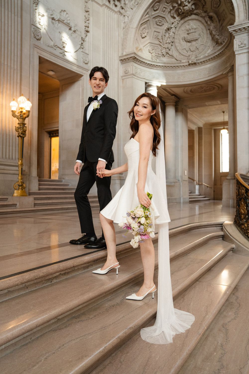 A wedding couple poses playfully on elegant marble stairs in a grand historic building with ornate architectural details.