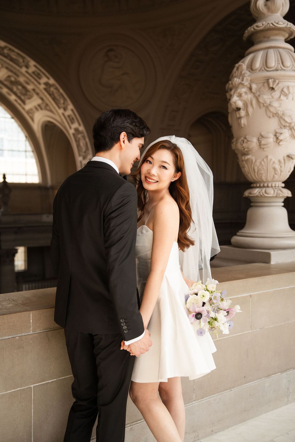 A romantic wedding couple poses in an elegant architectural setting with ornate stone columns and arches.