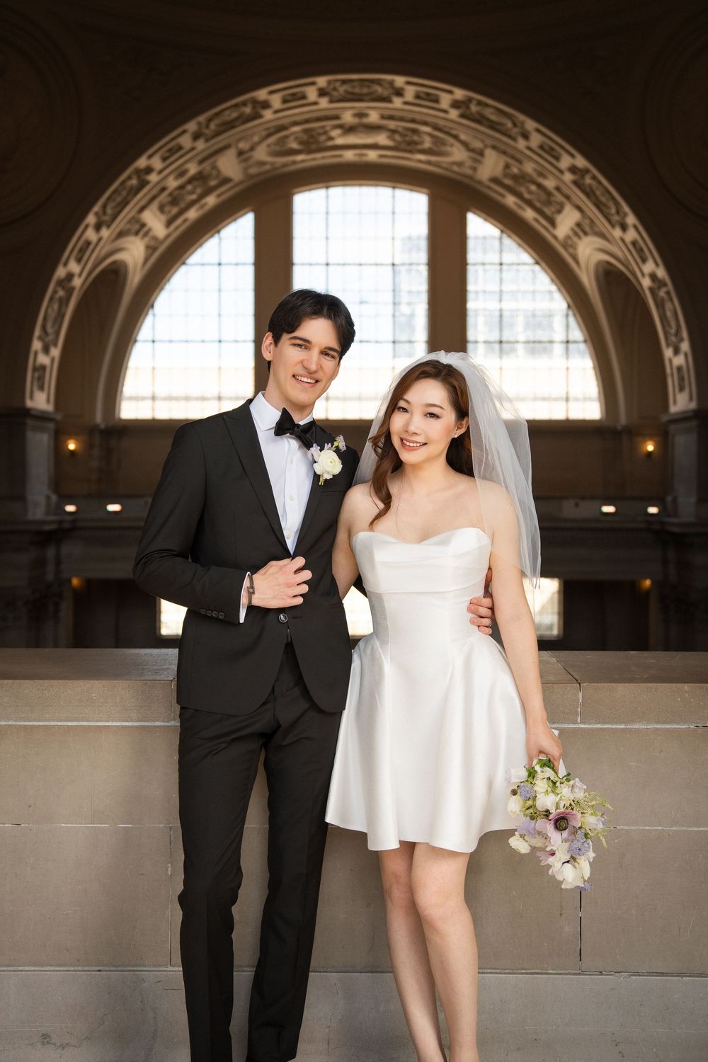 A couple in wedding attire poses under an ornate arched doorway with decorative stonework and a window.