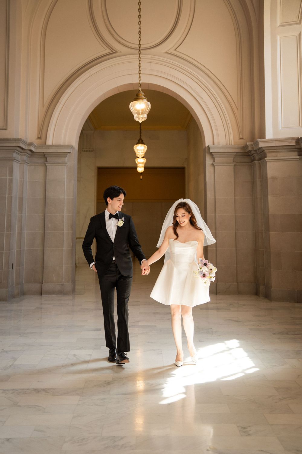 A couple in wedding attire dances joyfully across a sunlit marble hallway with classical architecture.