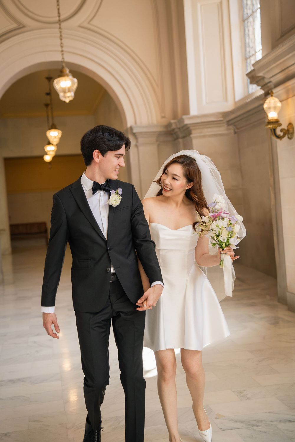 Couple in wedding attire walking and laughing together in an elegant hallway with arched ceilings and hanging lights.