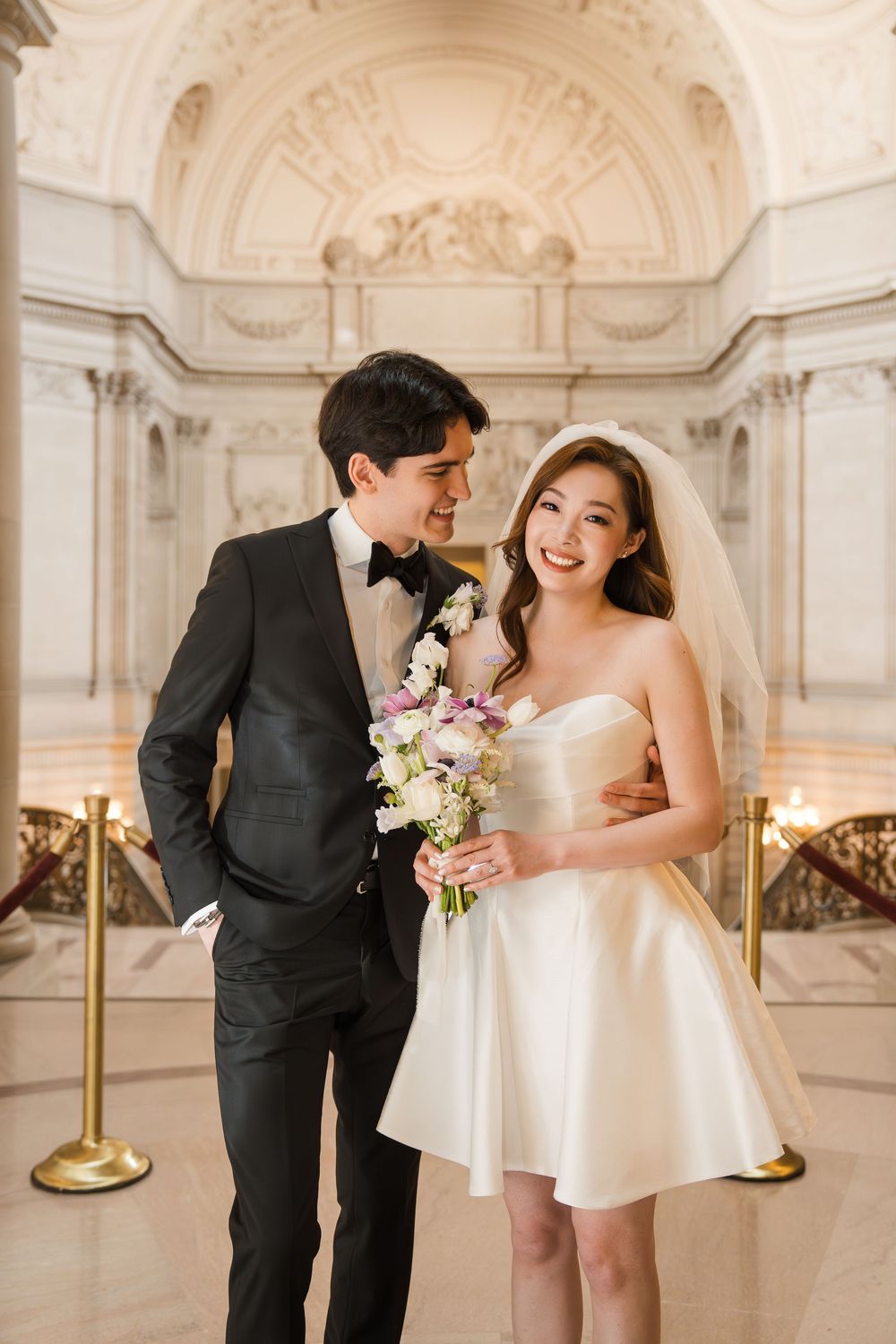 A couple in formal wedding attire share a joyful moment at San Francisco City Hall with ornate architecture in background.