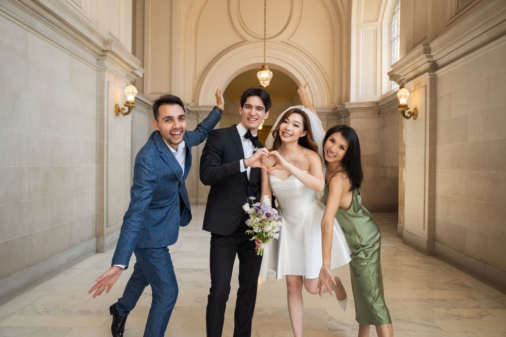 A wedding party poses playfully in a grand marble hallway with elegant architectural details and hanging light fixtures.