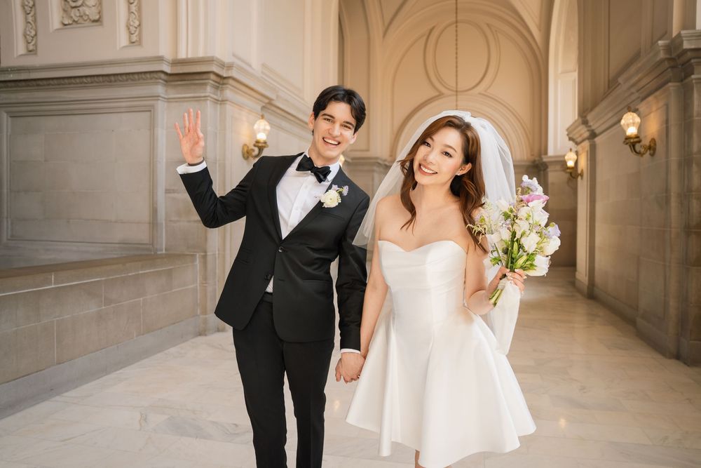 A joyful couple in wedding attire laughs and celebrates in an elegant marble hallway with ornate architecture.