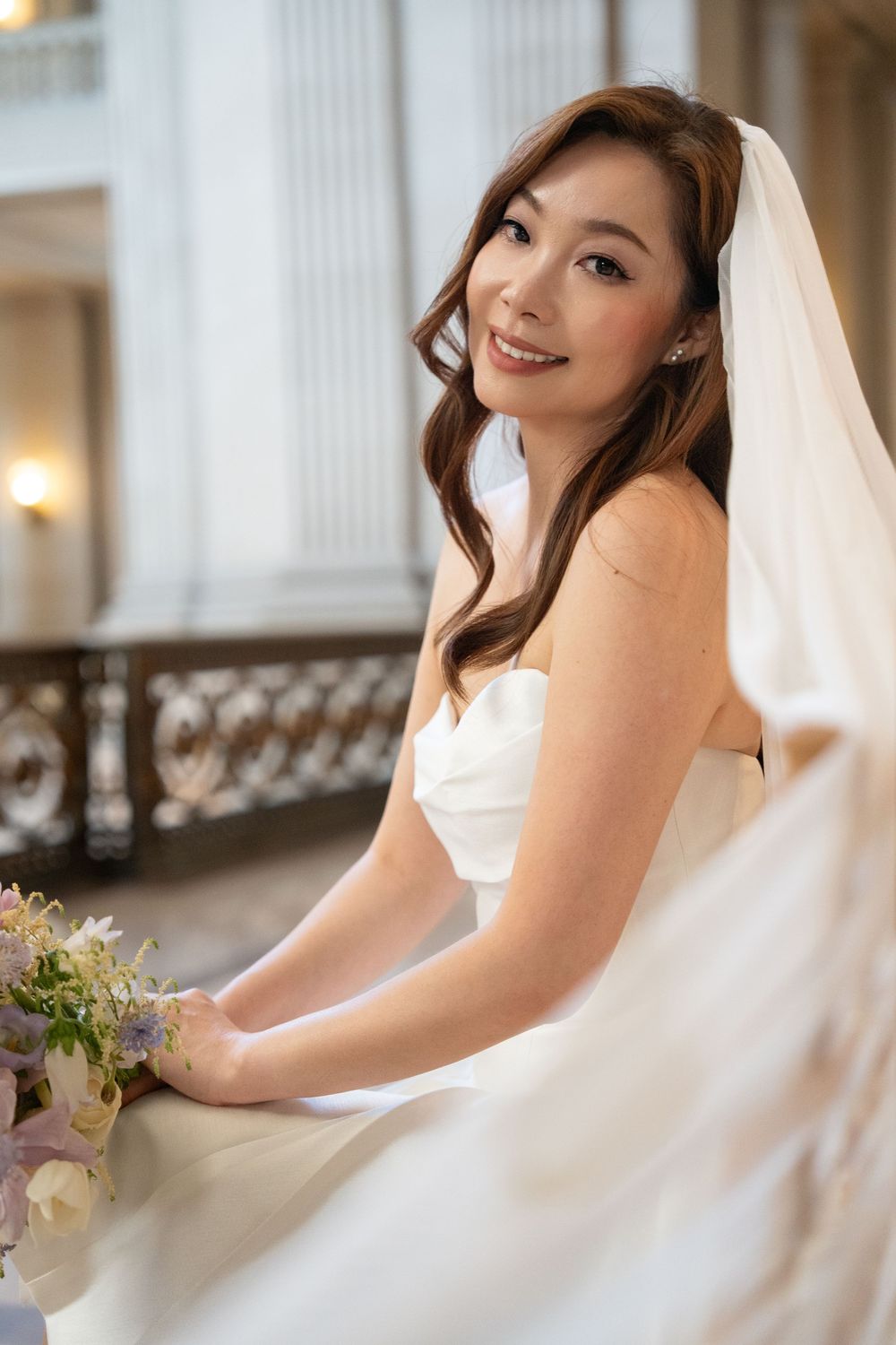 A bride in a white wedding dress poses elegantly with a bouquet in an indoor setting with soft lighting.