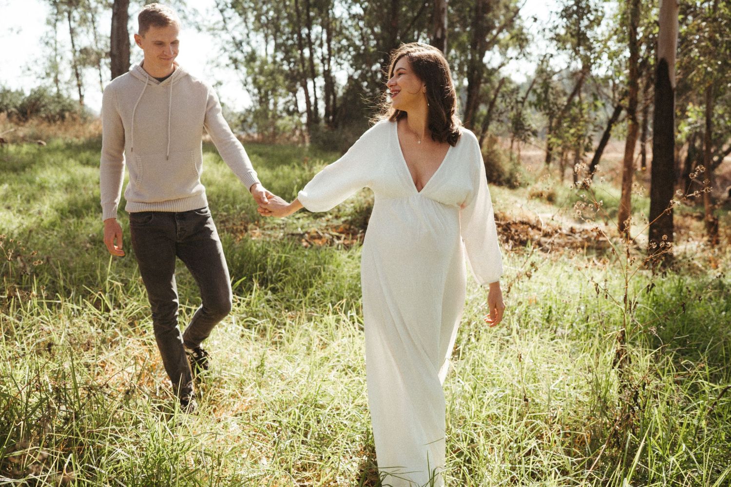 Couple in white dress and casual attire walking through sunny woodland setting.