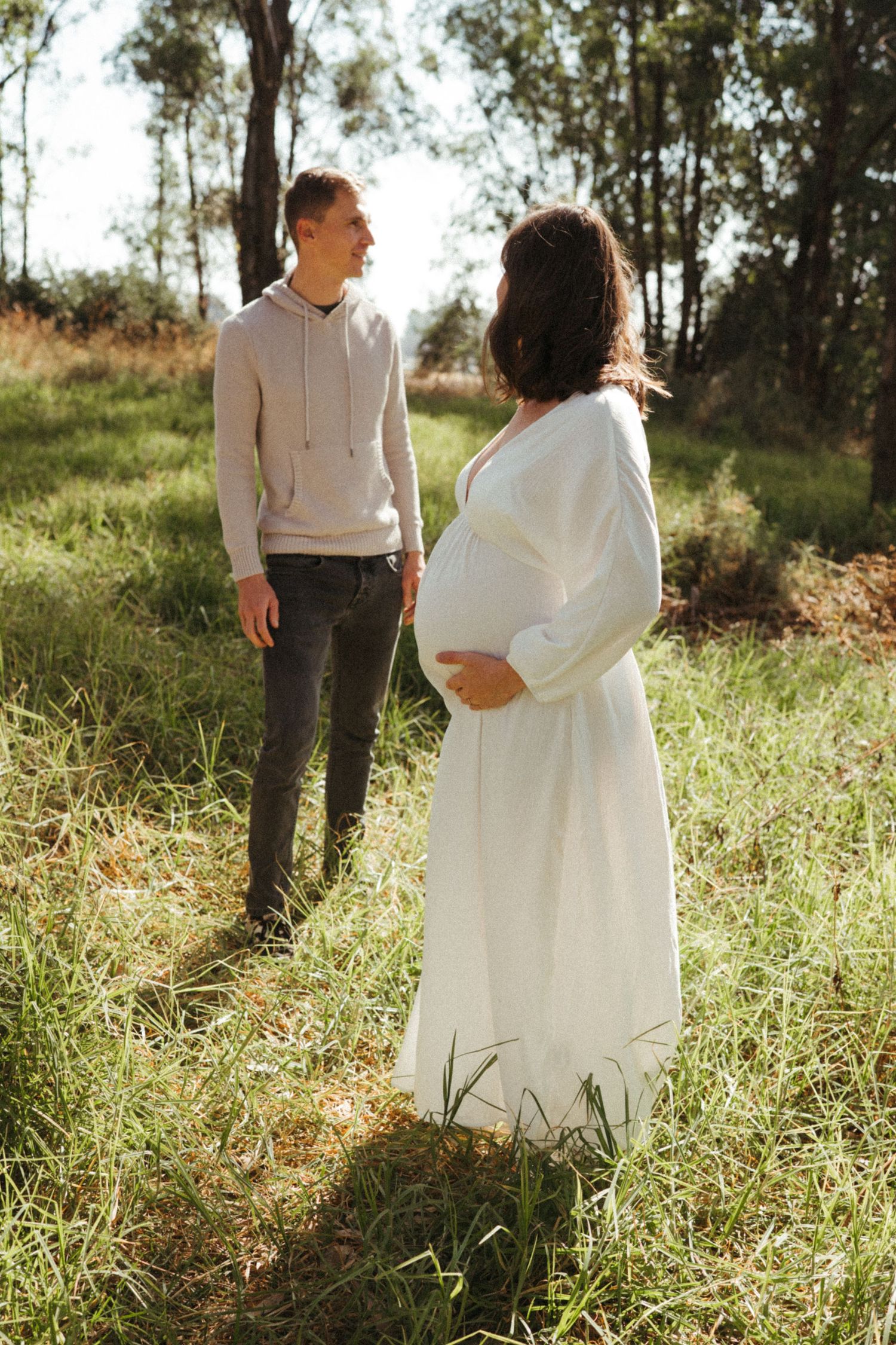 A couple stands together in tall grass with sunlight filtering through trees in the background.