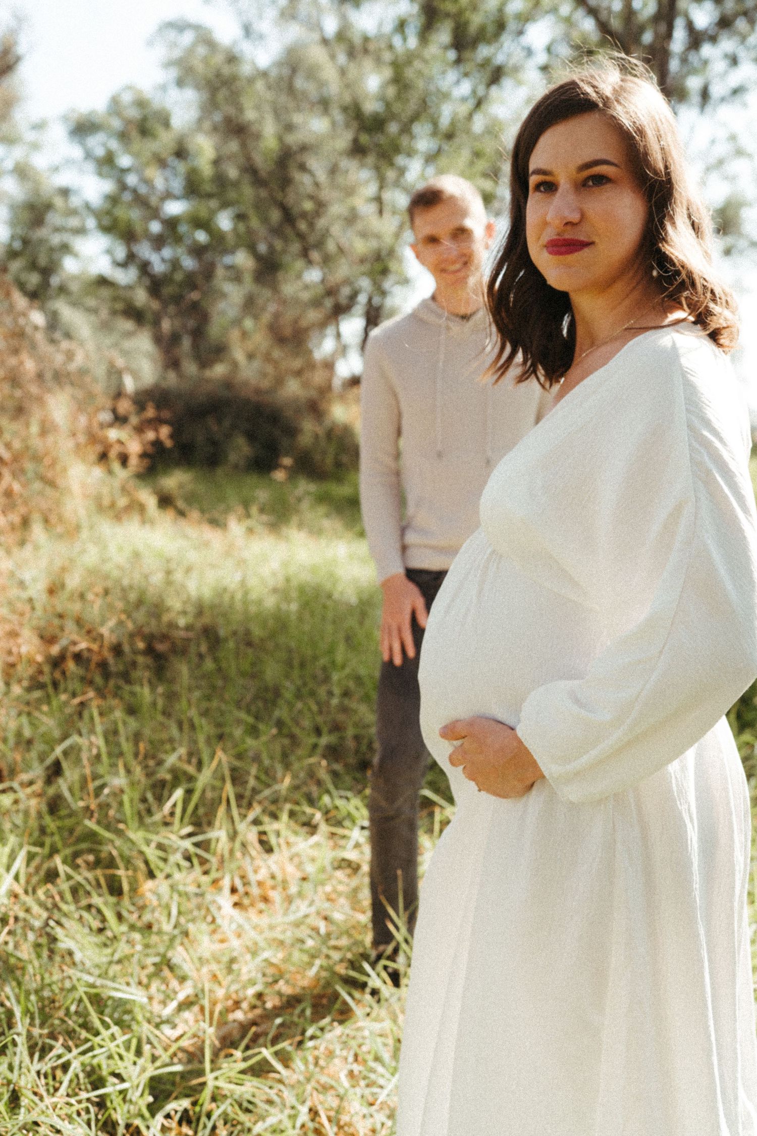 Sunlit outdoor maternity photoshoot in a field with flowing white dress.
