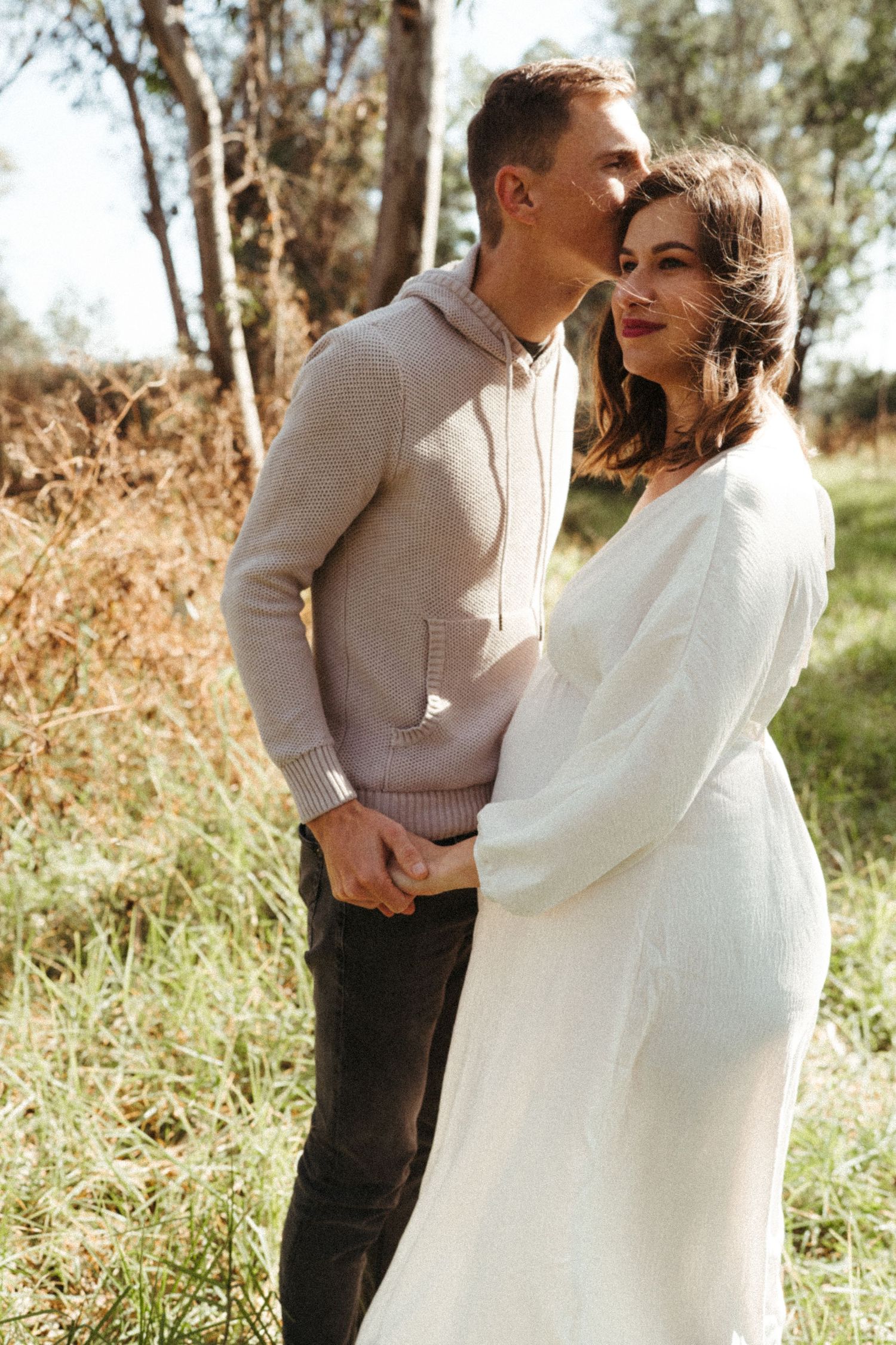 A couple shares an intimate moment in a field, wearing a beige sweater and flowing white dress.