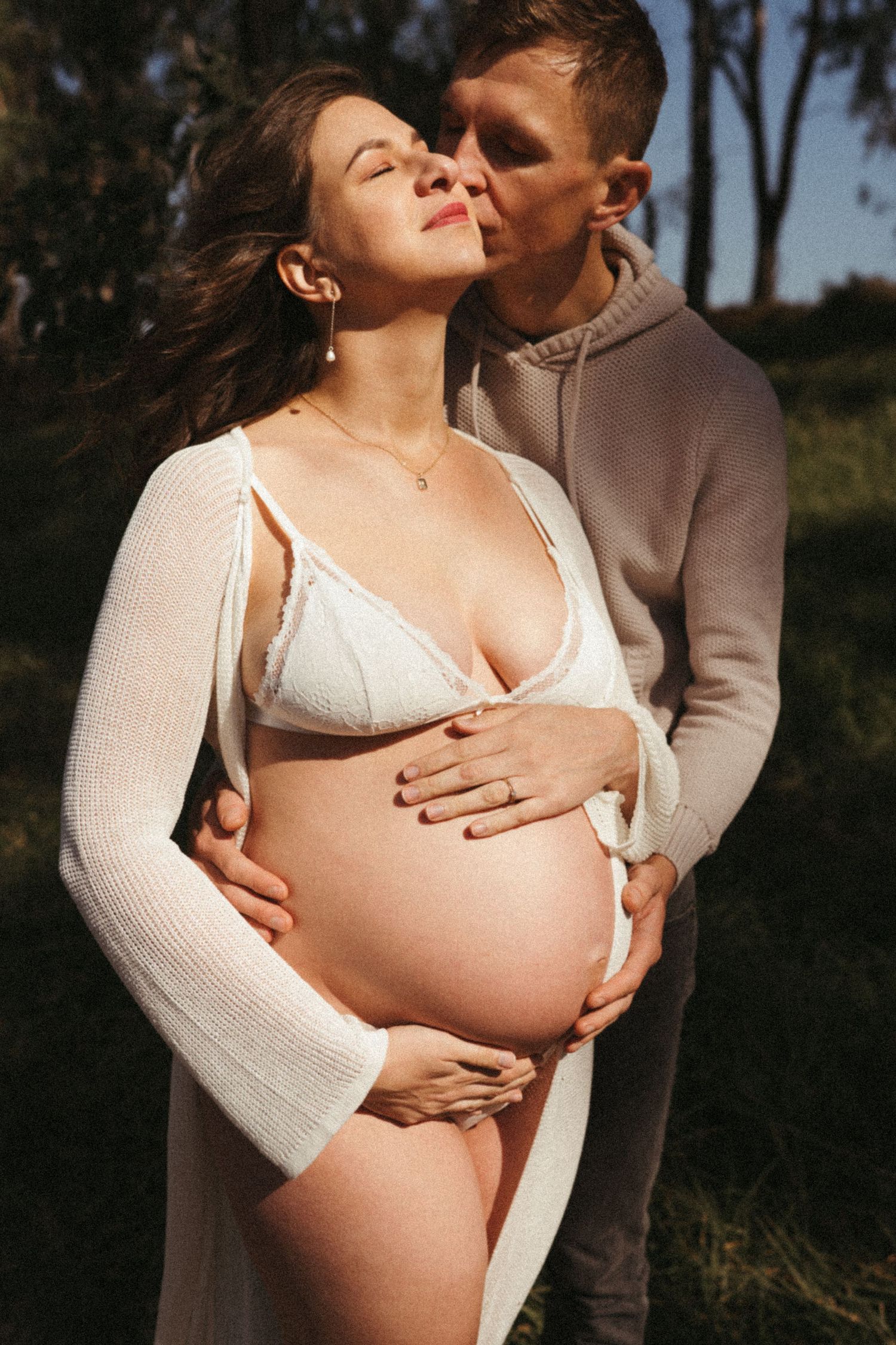 Intimate maternity photoshoot in white lingerie and cardigan against a natural outdoor backdrop.