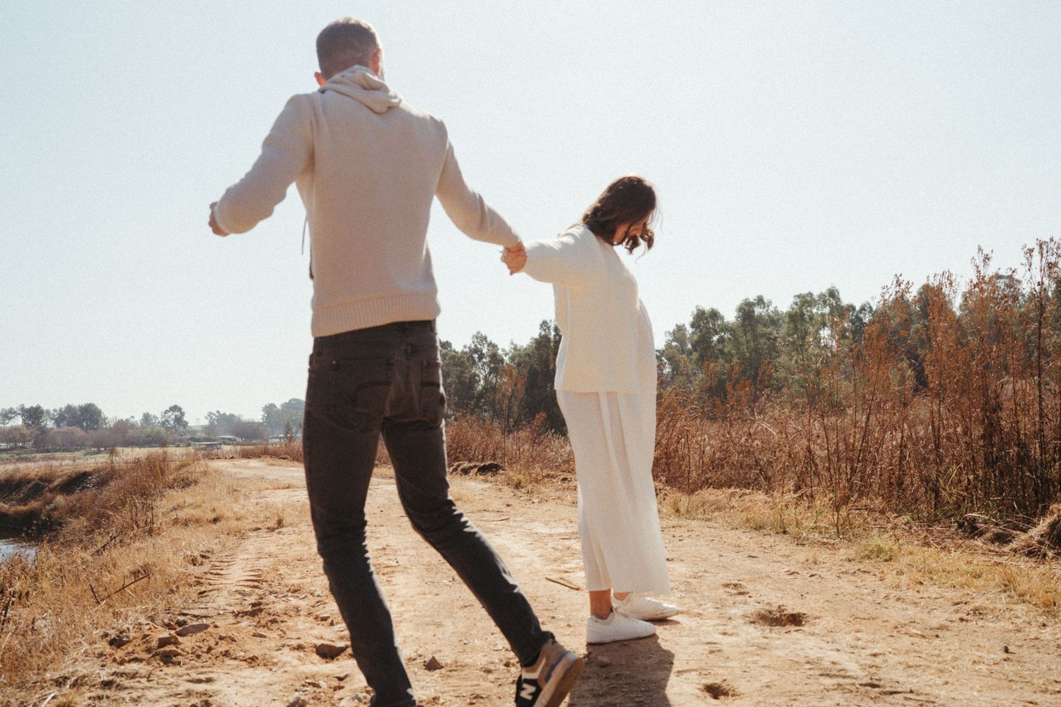 Couple walking hand in hand on a sunlit autumn path during maternity shoot.