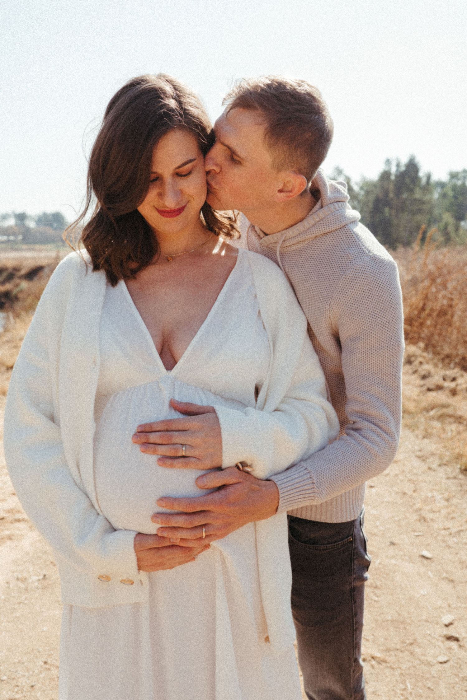 Tender maternity moment captured in warm sunlight with white sweater.