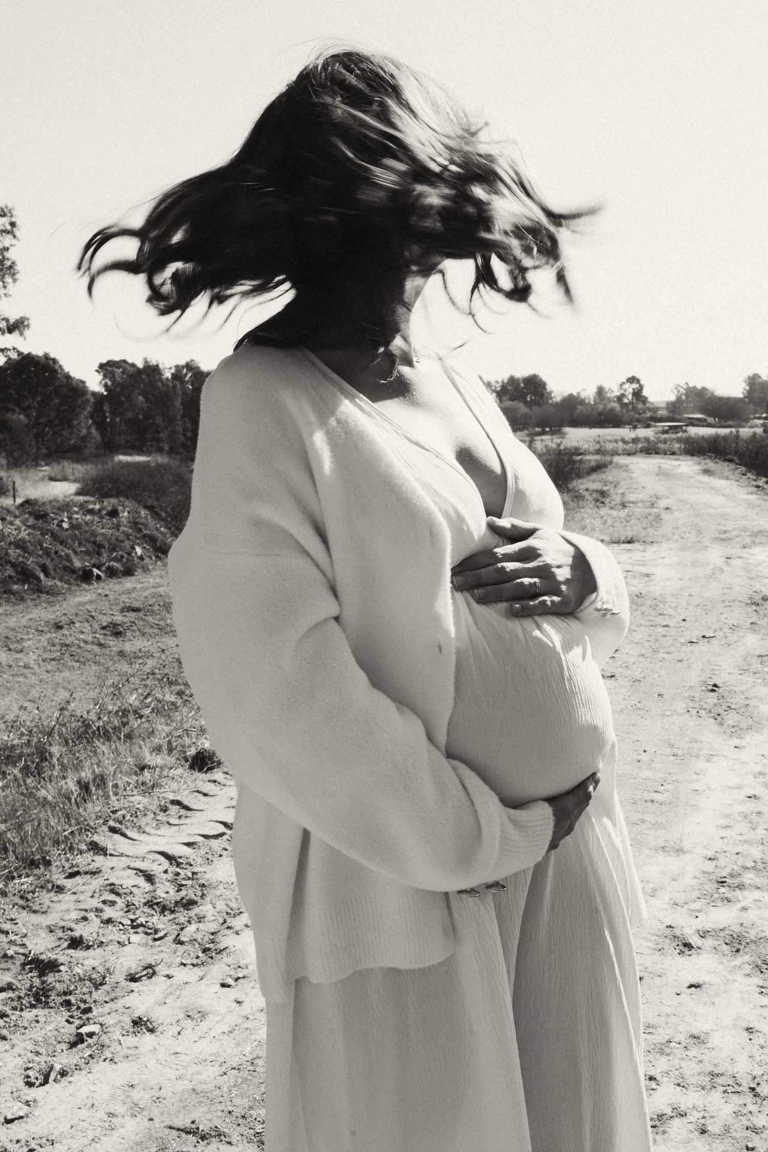 A silhouetted figure in a flowing white dress stands on a dirt path with windswept hair.