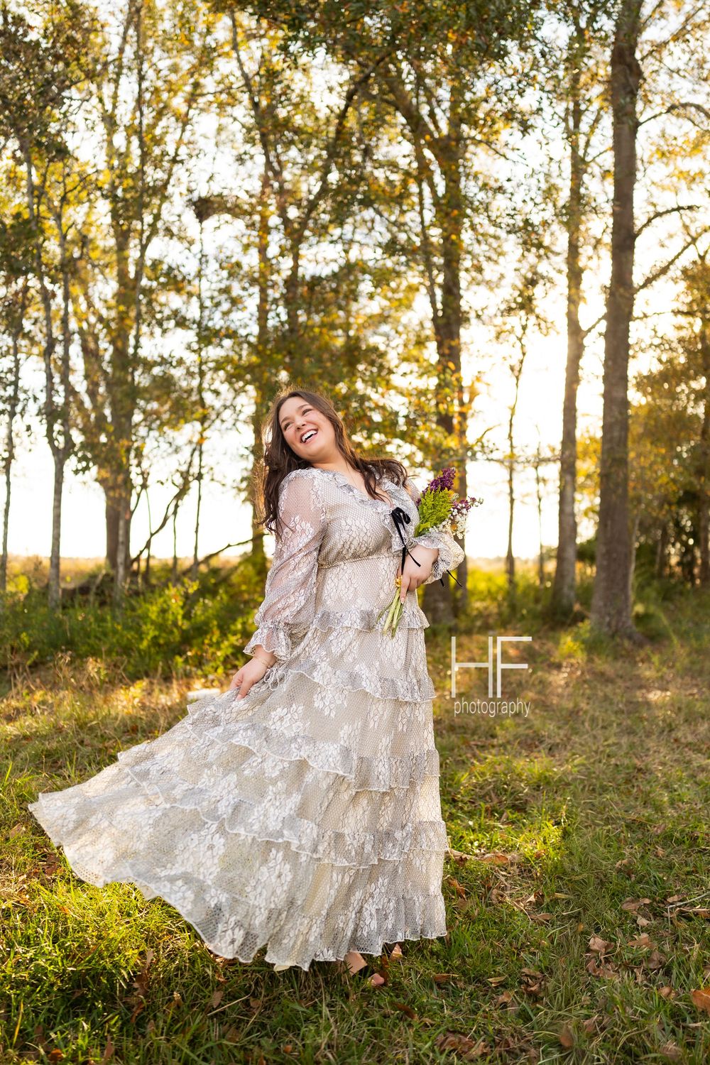 A joyful outdoor portrait featuring a layered lace dress and purple bouquet in dappled sunlight.