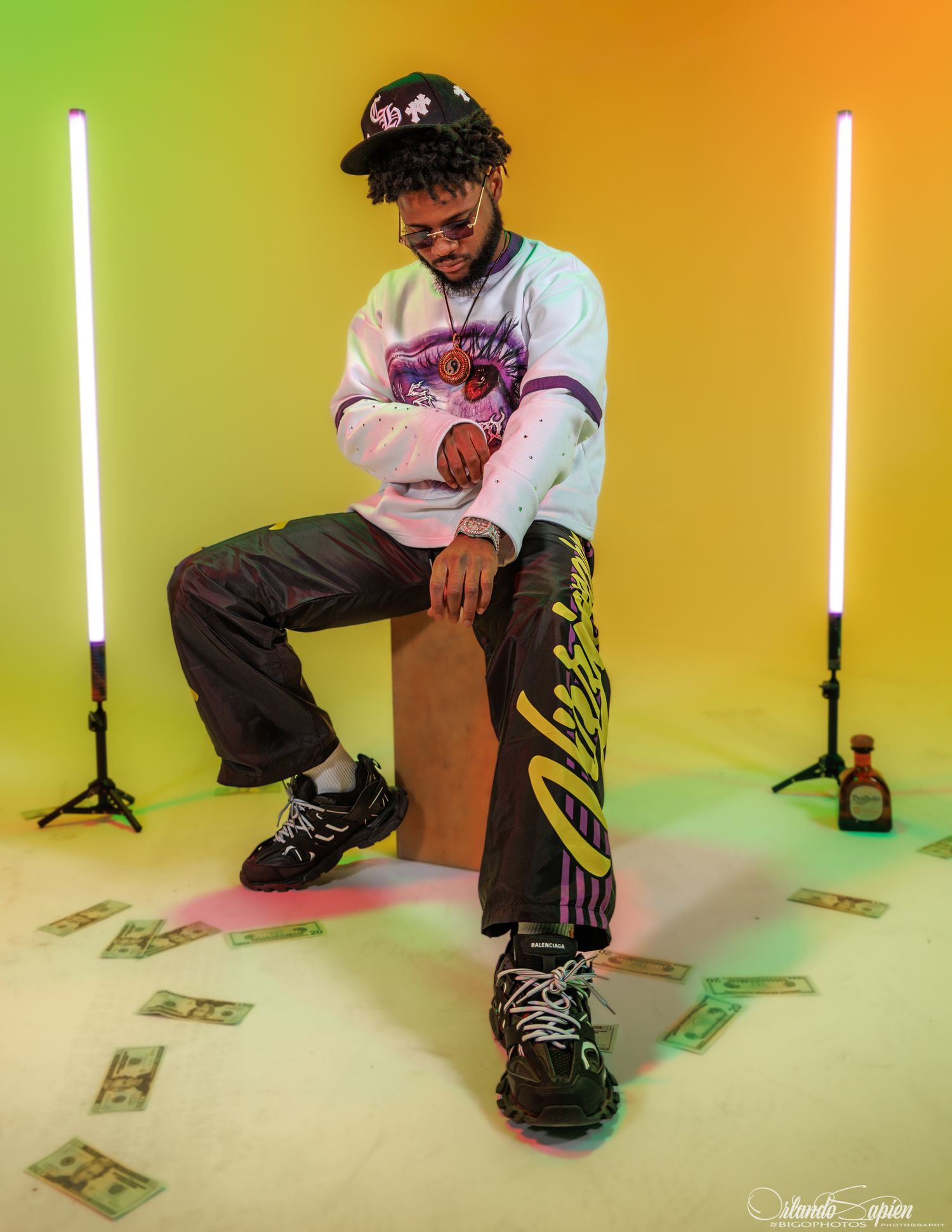 Person in baggy clothes sits with dollar bills scattered on floor against colorful gradient background with vertical lights.