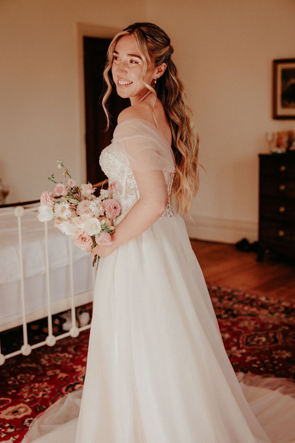 A wedding dress with off-shoulder design and long flowing train is showcased in a warm-lit room with a delicate bouquet.