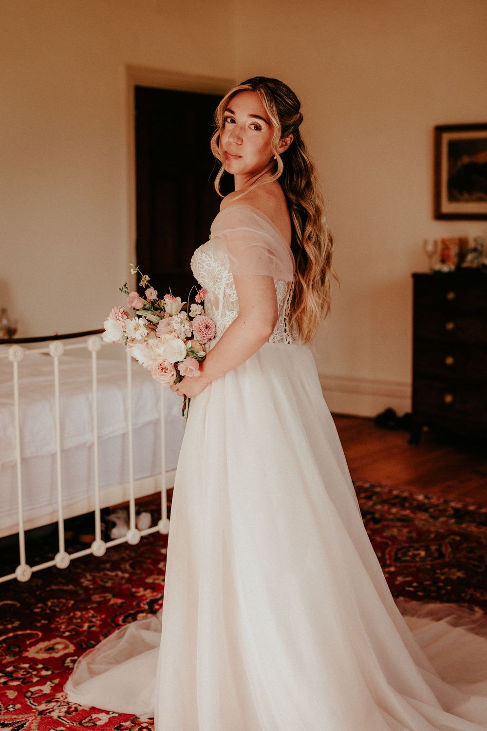 A bride in a flowing white lace wedding gown poses gracefully against a warm-toned interior setting with rose petals on the floor.