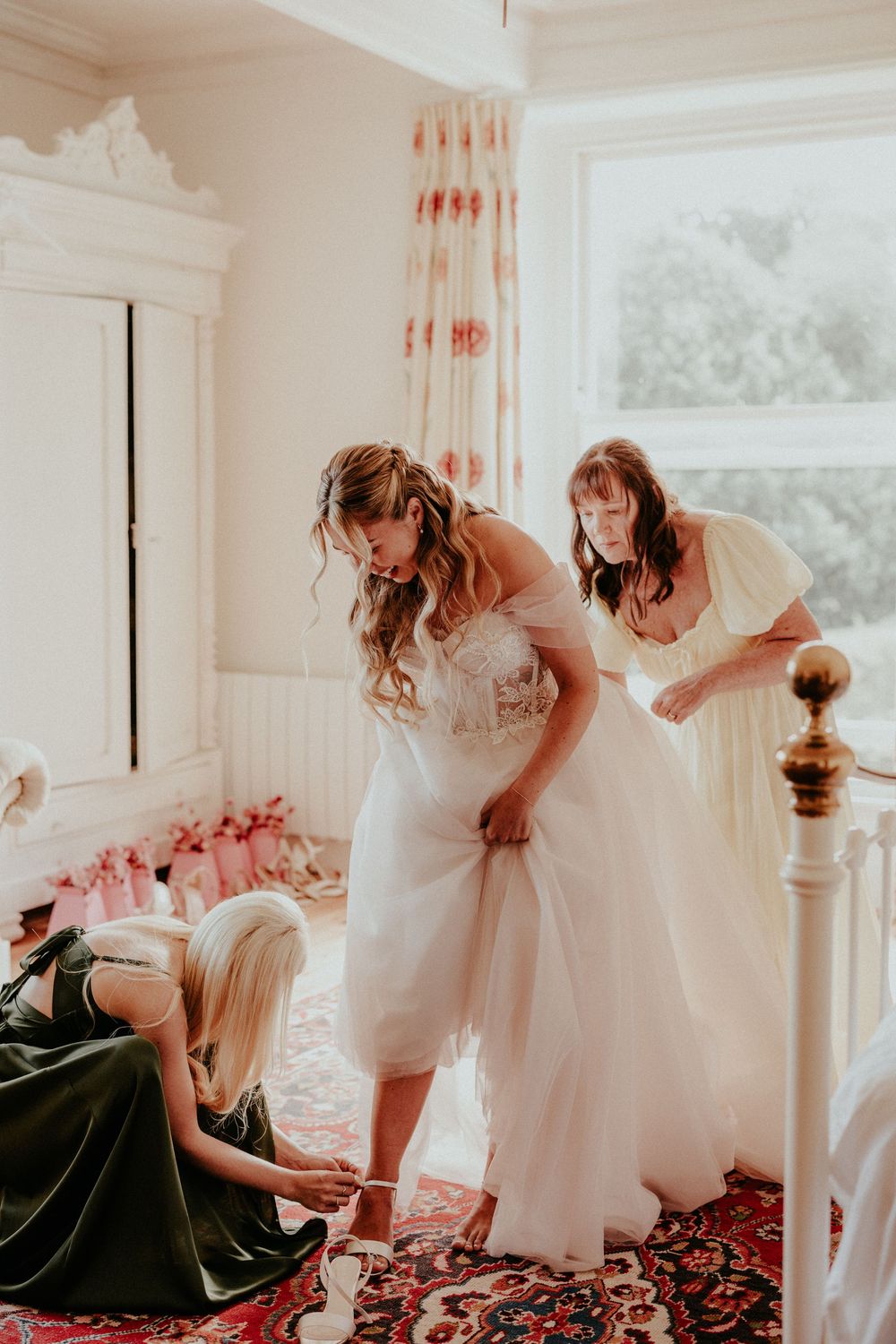 Wedding dress preparation scene with helpers assisting in a sunlit room with vintage decor and red carpet.
