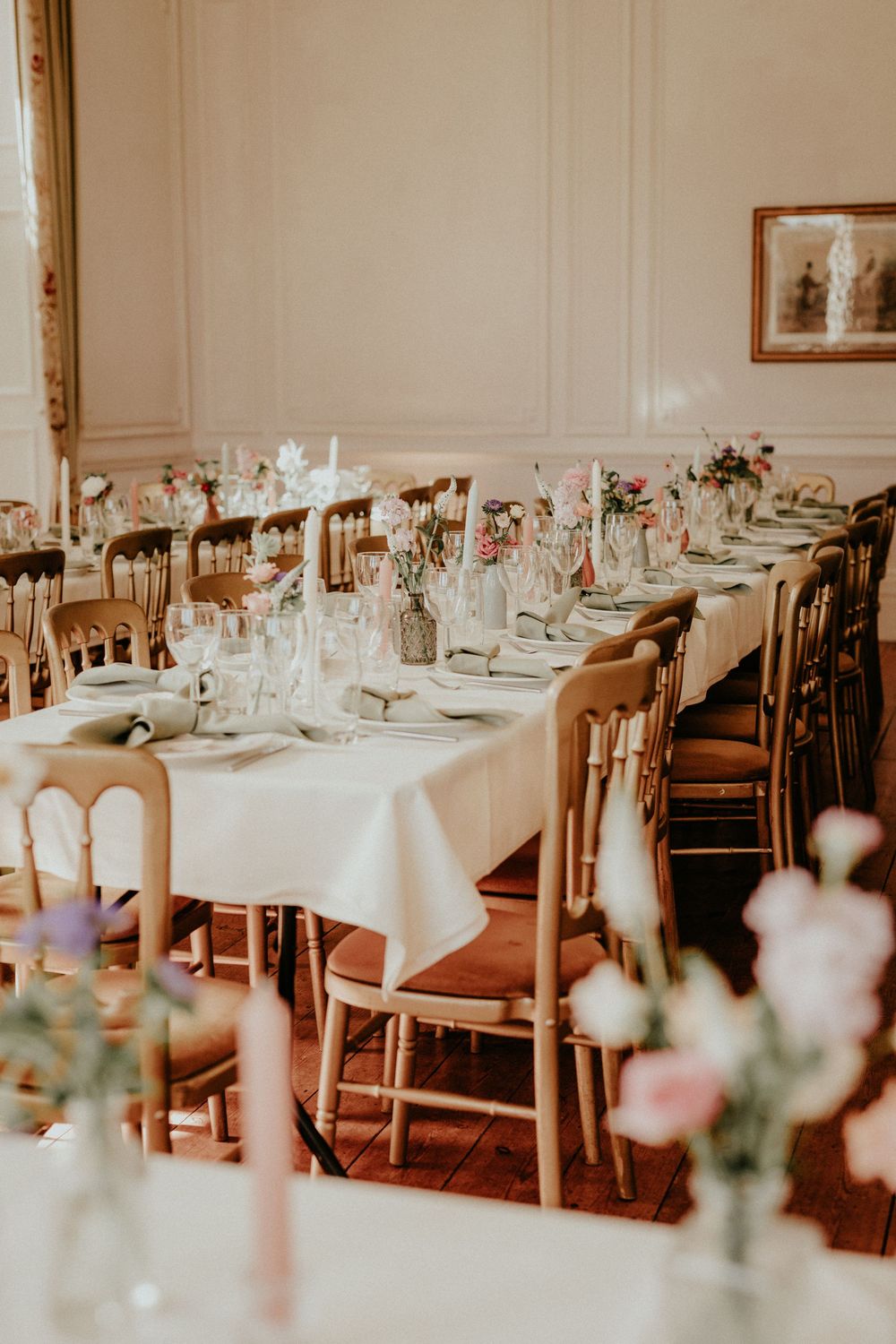 Elegant wedding reception tables set with white linens, floral centerpieces and wooden chairs in a bright banquet room.