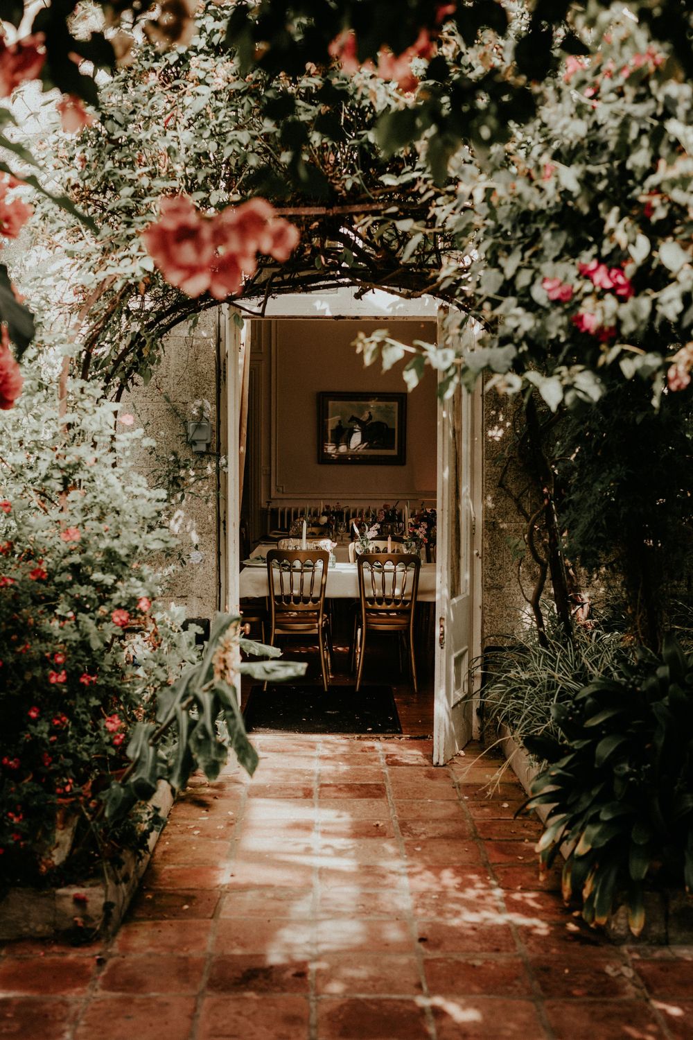 A romantic garden pathway leads to an open doorway revealing a cozy dining room with wooden chairs and soft lighting.