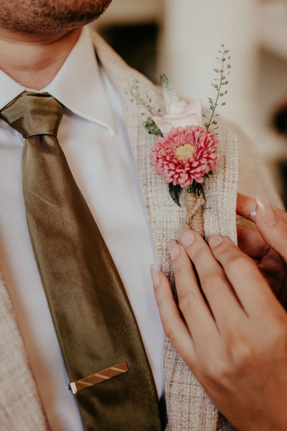 Hands pinning a pink carnation and white floral boutonniere onto a brown tie and white dress shirt.