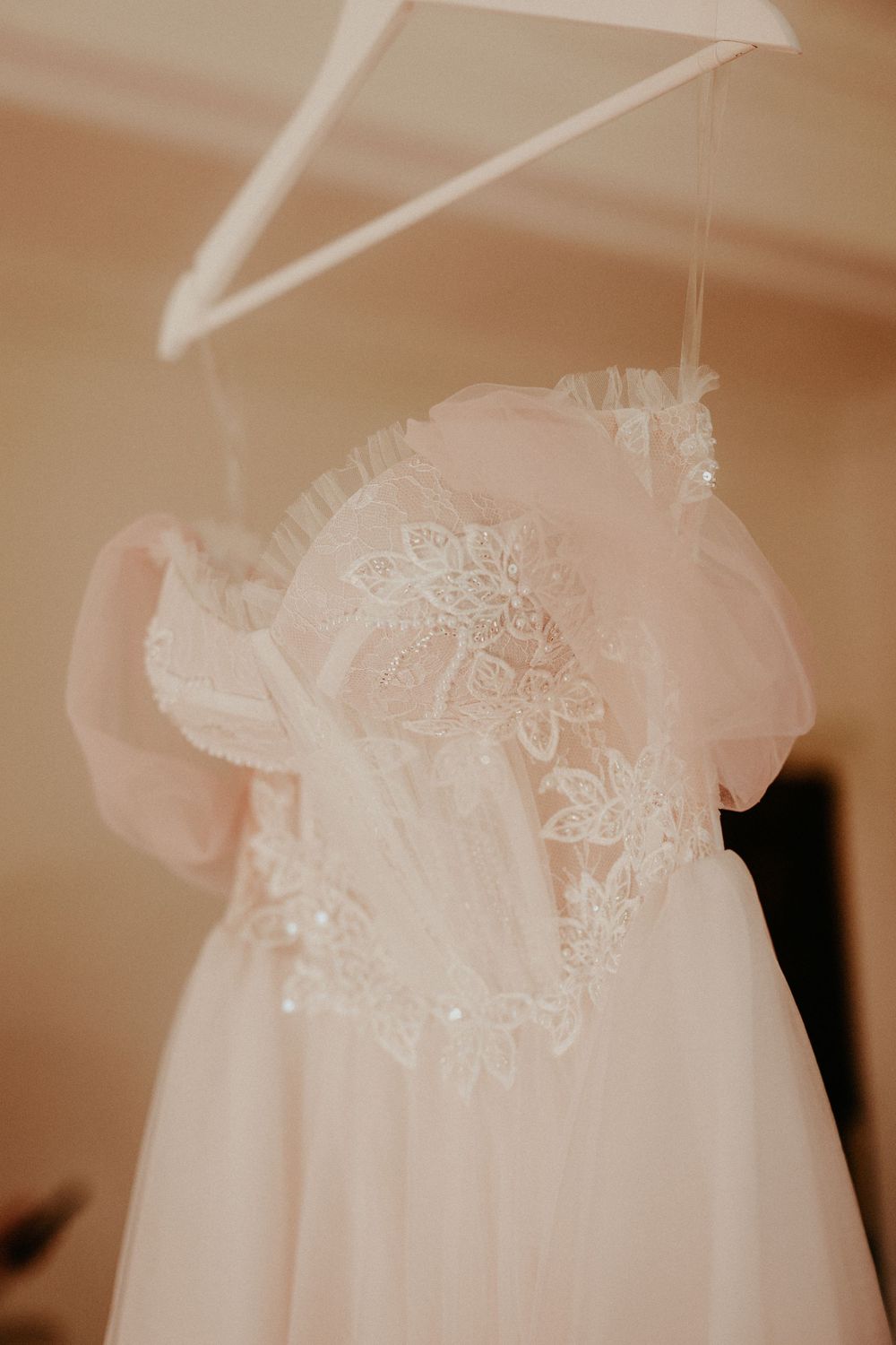 A delicate white wedding dress with lace details and tulle fabric hangs on a white hanger against a neutral background.