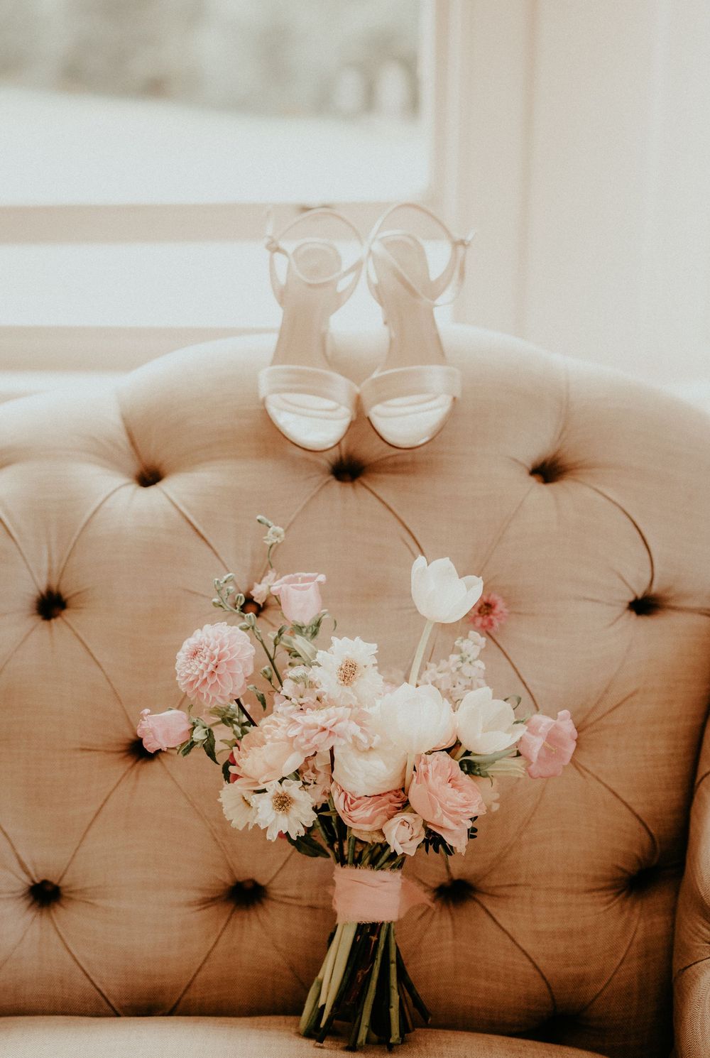 White bridal shoes and a soft pink floral bouquet arranged on a tufted beige couch in natural light.