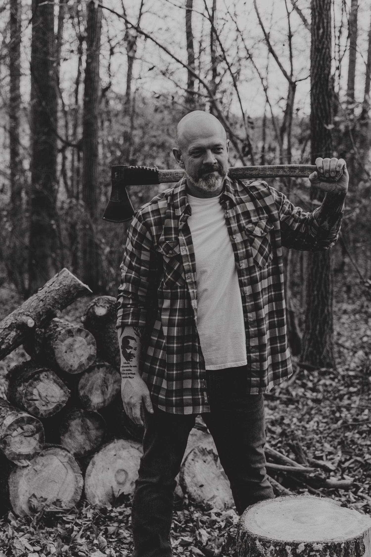 A person in a plaid flannel shirt carries an axe while standing next to a woodpile in a forest setting.
