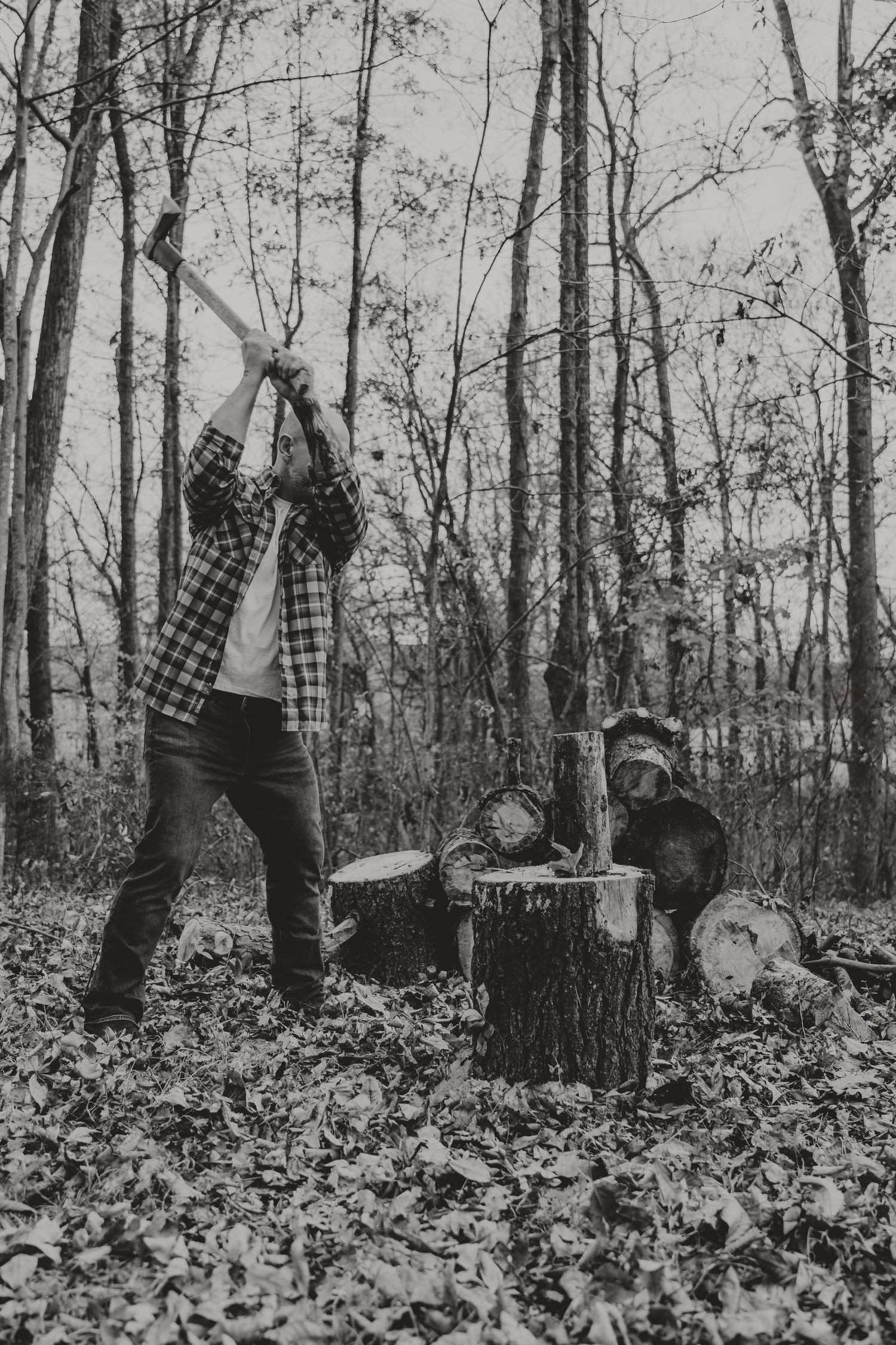 Black and white photo of someone chopping wood in a forest with an axe while fallen leaves cover the ground.