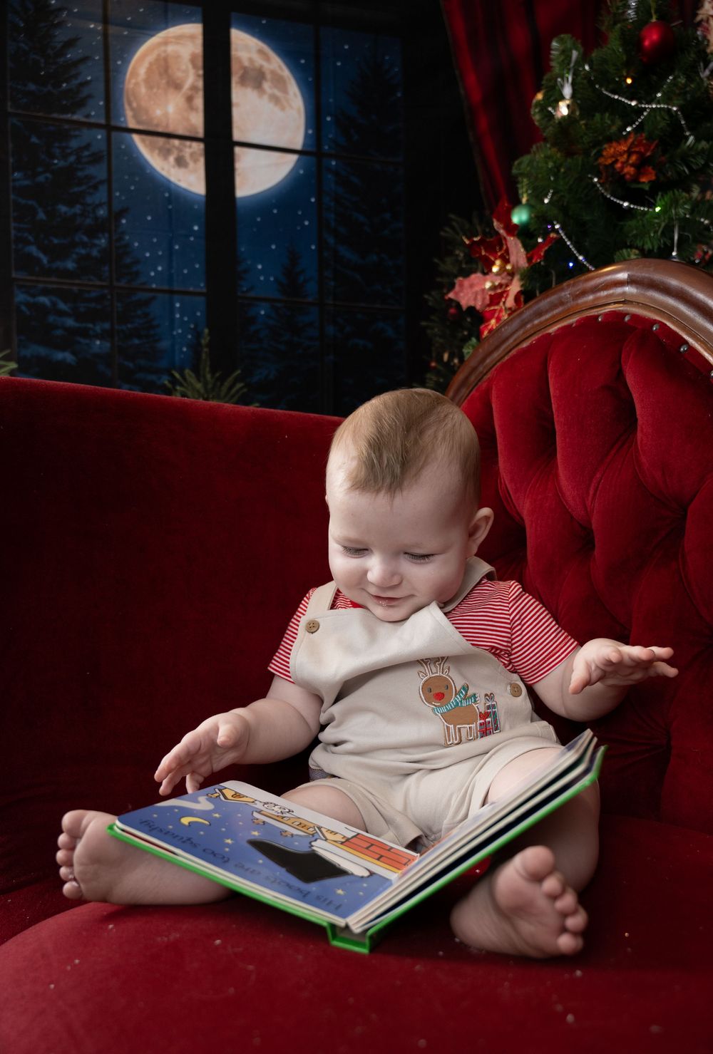 Baby in white onesie lounges on red velvet couch while reading a children's book in a festive holiday setting.