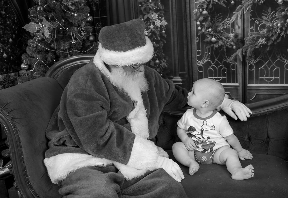 A vintage black and white photo of Santa Claus sitting in a chair with a small baby in holiday attire.