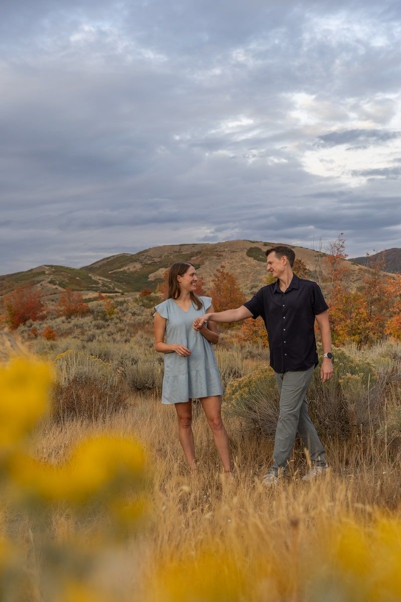 A couple shares a tender moment in a golden autumn field with mountains and cloudy skies in the background.