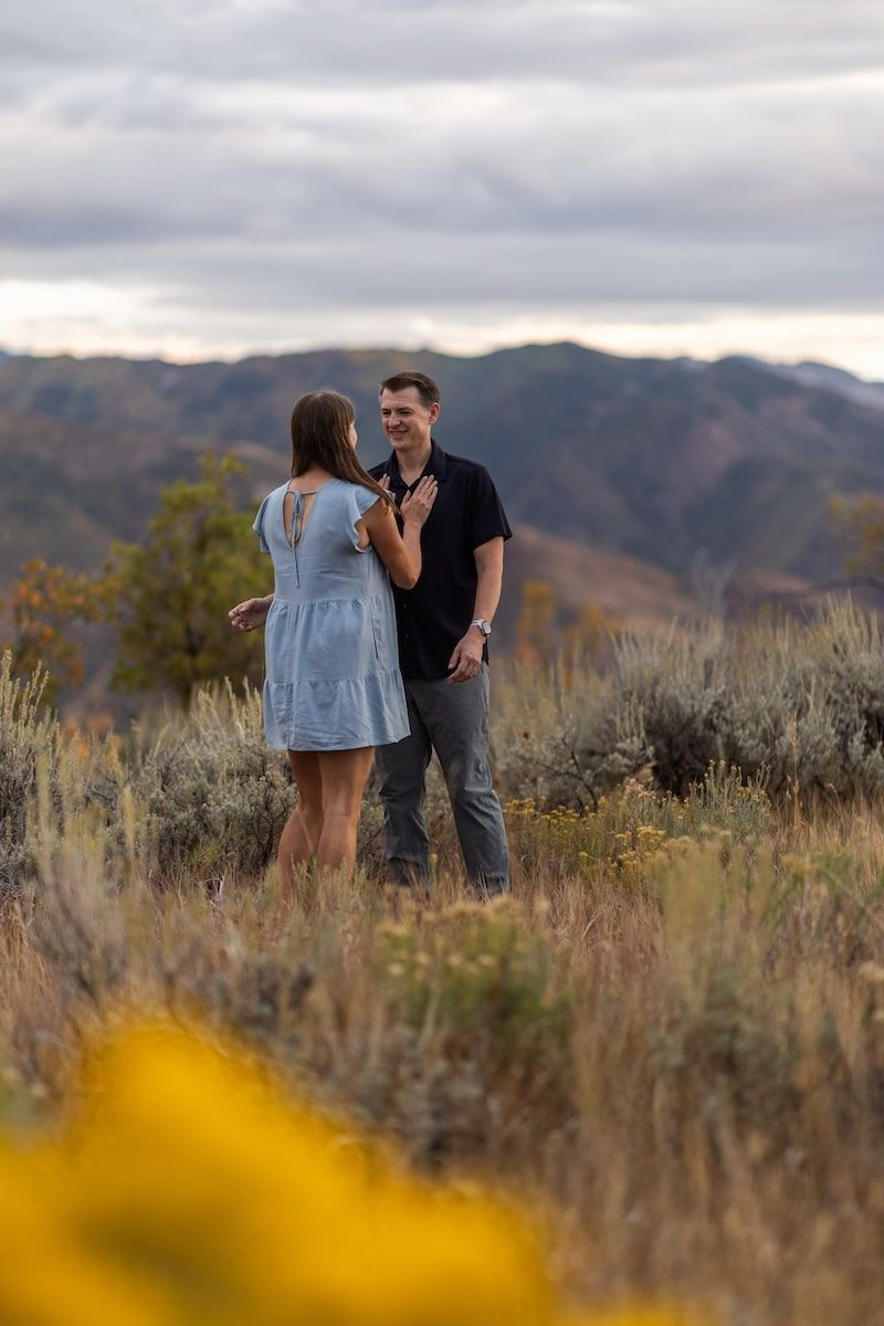 A couple shares a romantic moment in a field of tall grass with mountains in the background at sunset.
