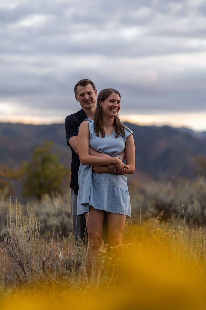 A couple embraces in a desert landscape with mountains and sagebrush at sunset.