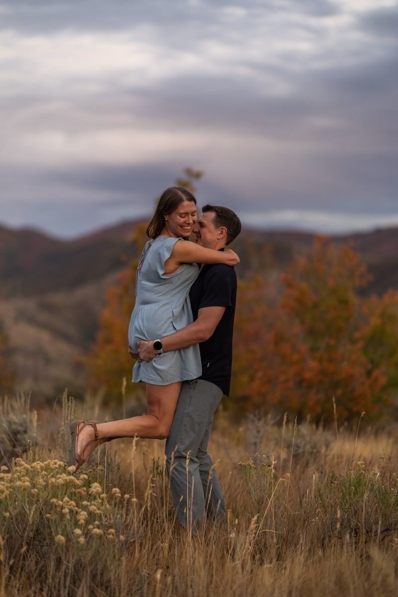 A couple embraces in a romantic moment against a backdrop of mountains and autumn foliage at sunset.
