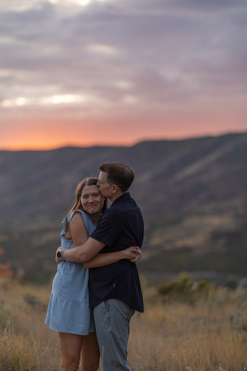 A couple embraces in a romantic moment at sunset on a hilltop overlooking a mountainous landscape.