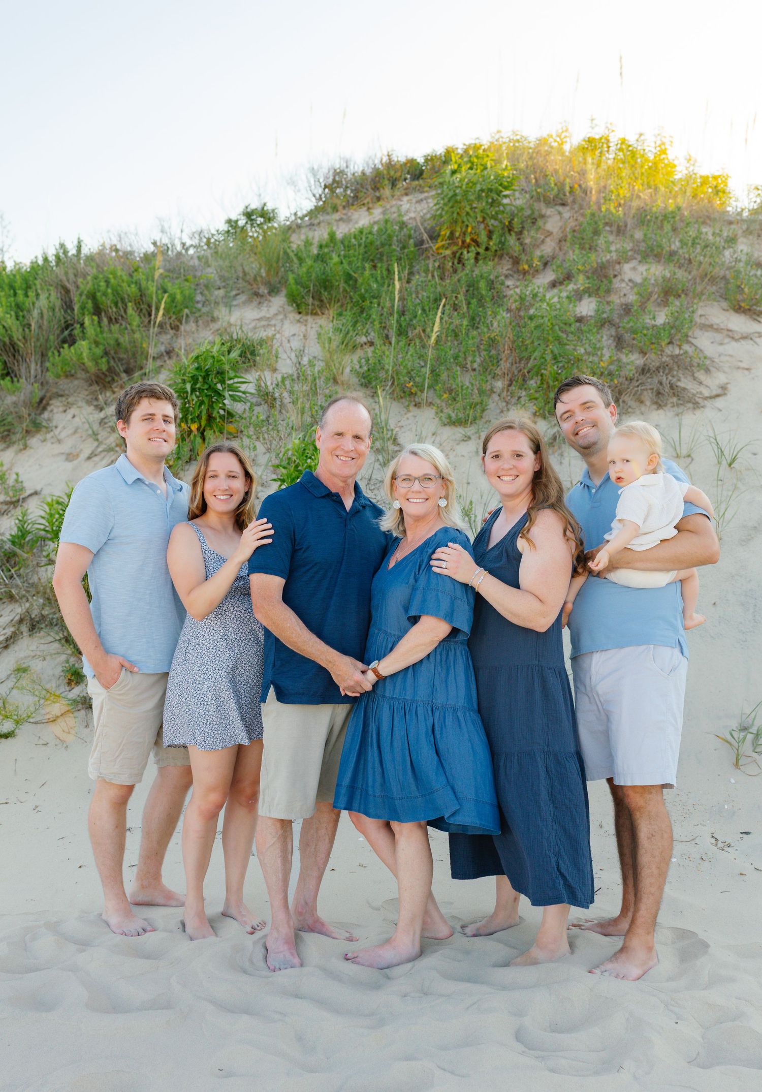 Family group poses together on sandy beach dunes at sunset with natural vegetation in background.