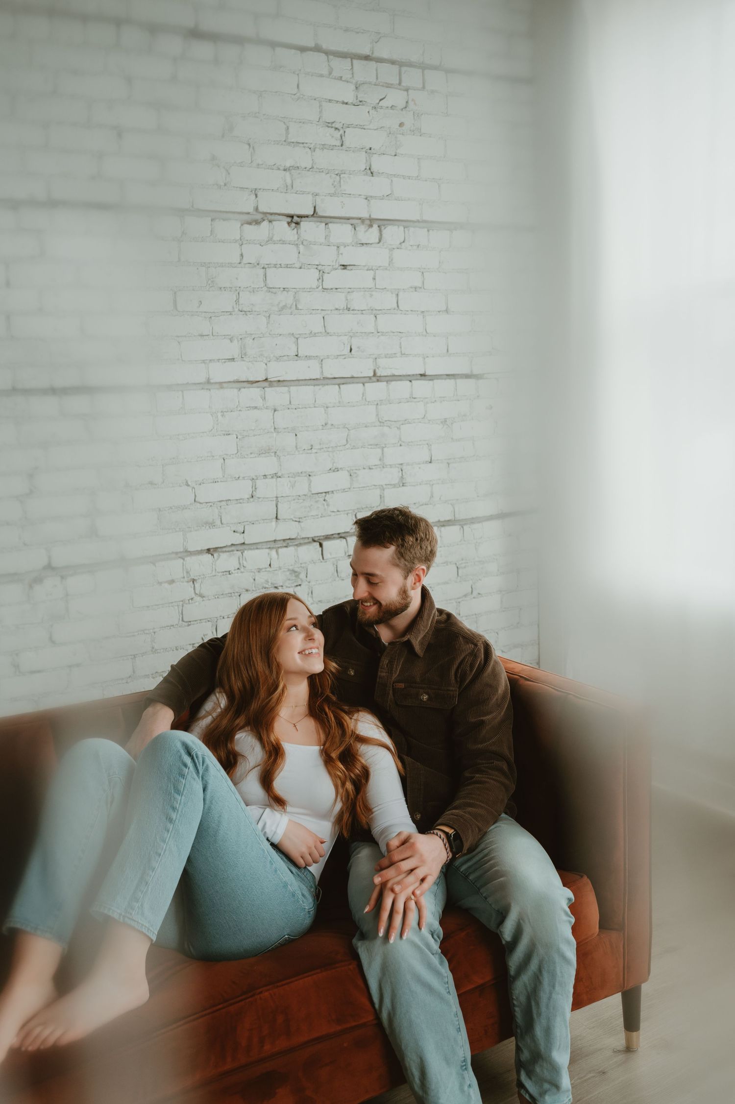 Romantic couple lounging together on a brown leather couch against white brick wall.