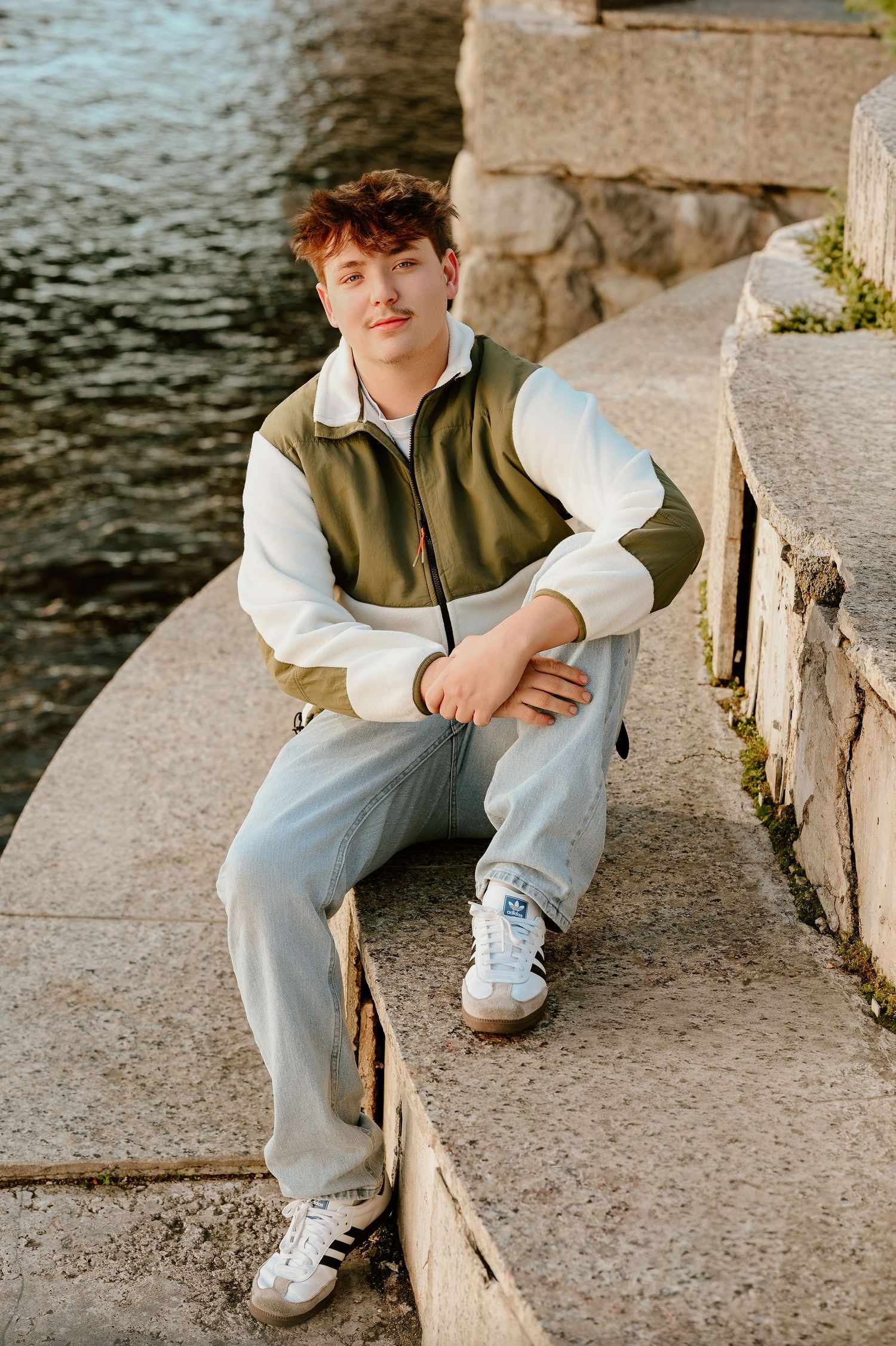 Someone wearing light jeans and white sneakers sits casually on stone steps near water on a sunny day.