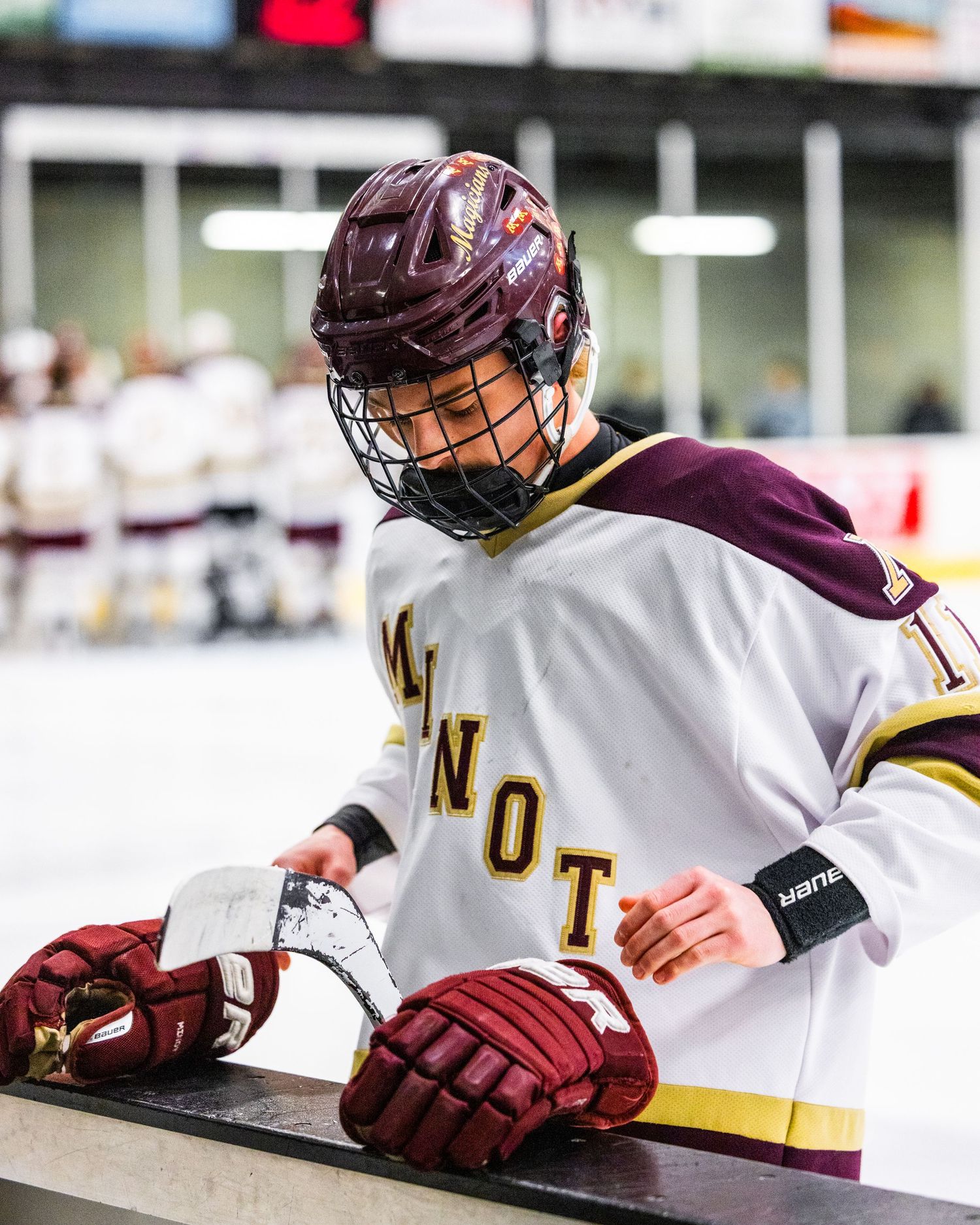 Hockey player in white jersey labeled 'NOT' adjusts their maroon gloves while standing at the bench.