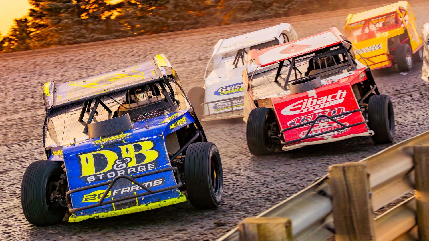 Race cars line up on a dirt track during a late model stock car racing event at sunset.