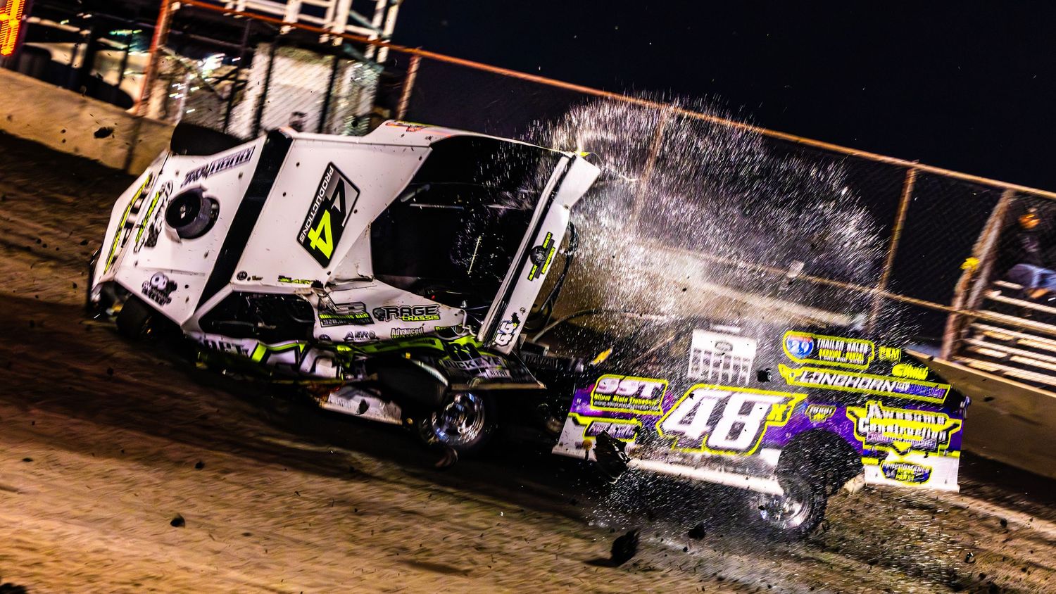Modified race car flips over during dramatic dirt track racing accident at night with bright lights illuminating the scene.