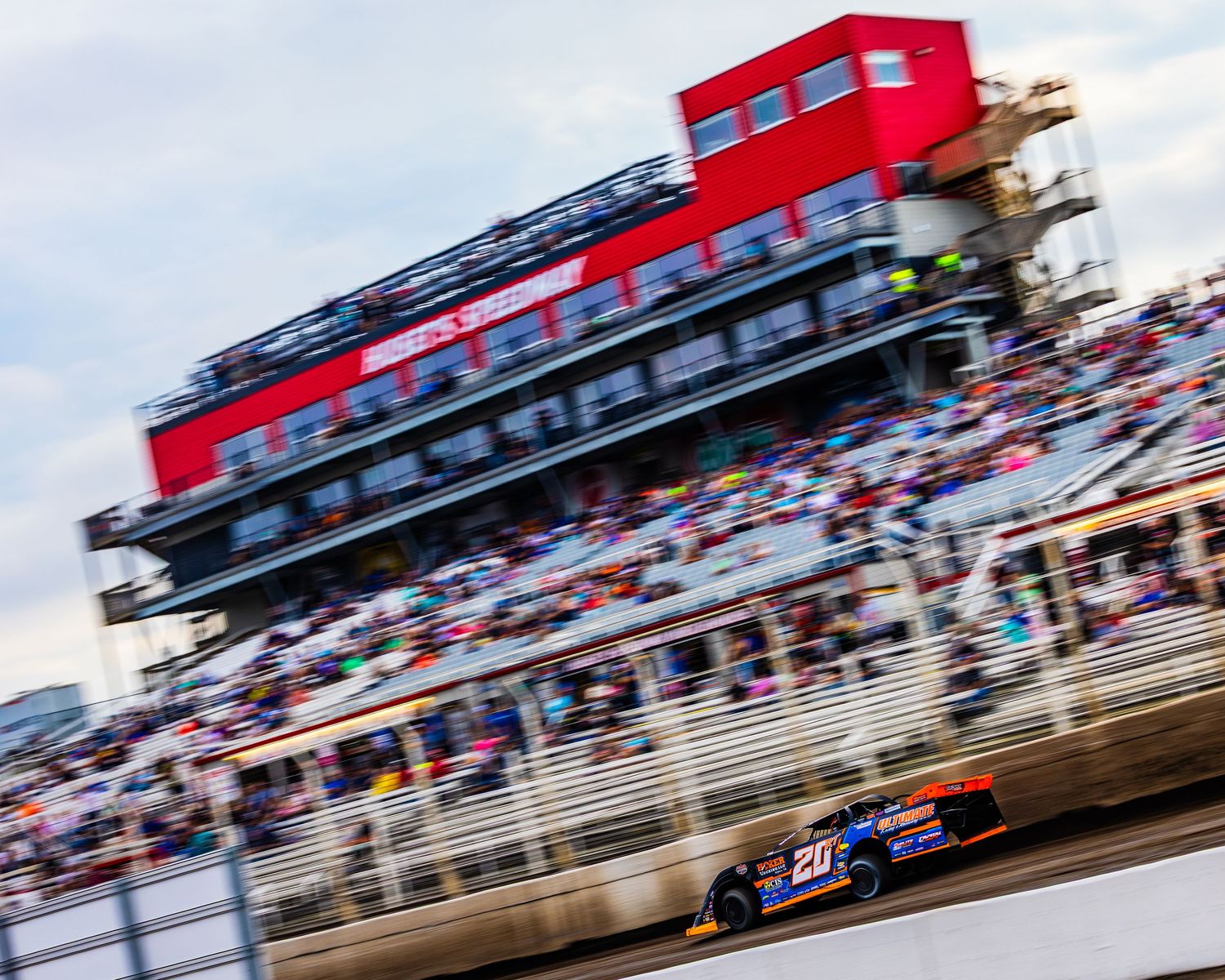 Red and white NASCAR race car speeds past crowded grandstands during a daytime racing event.