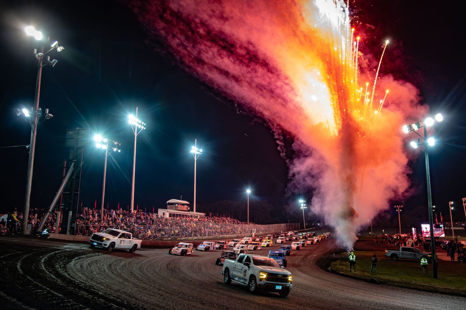 Red flares and bright stadium lights illuminate a nighttime racing event with cars on a dirt track.